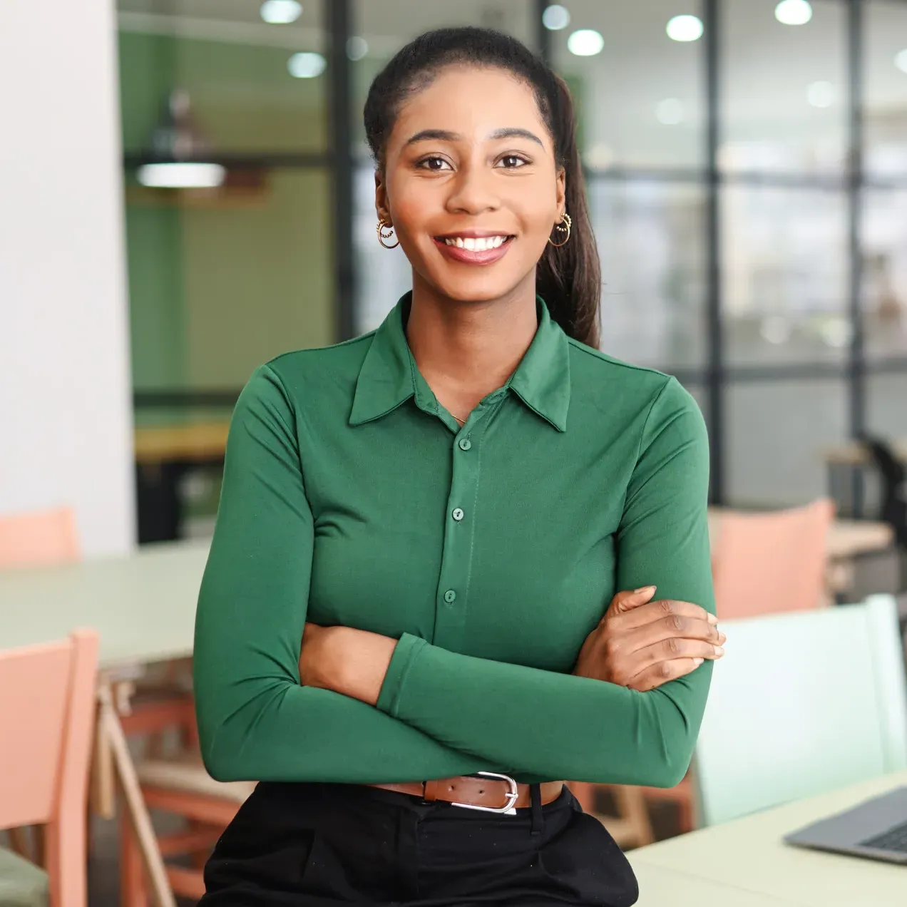 A professional woman in a green blouse sits at a modern office table, smiling with arms crossed, ready for career success.