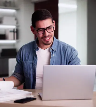 A man in a casual business setting studies financial documents and works on a laptop, representing career opportunities with a Herzing University accounting degree.